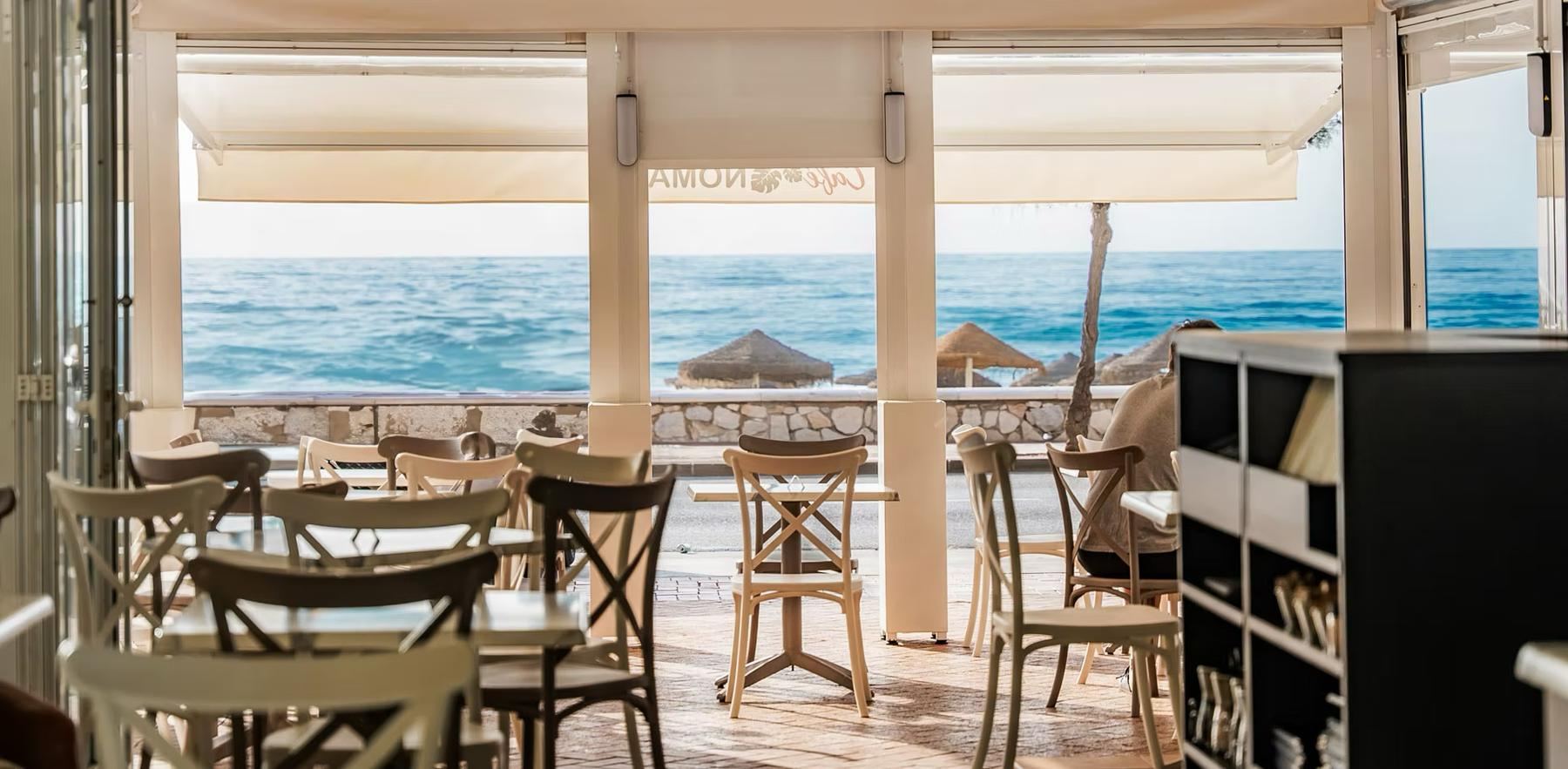 Der Gastraum zur Terrasse hin offen: Strohschirme, zwei Gäste an einem Tisch und das Mittelmeer.