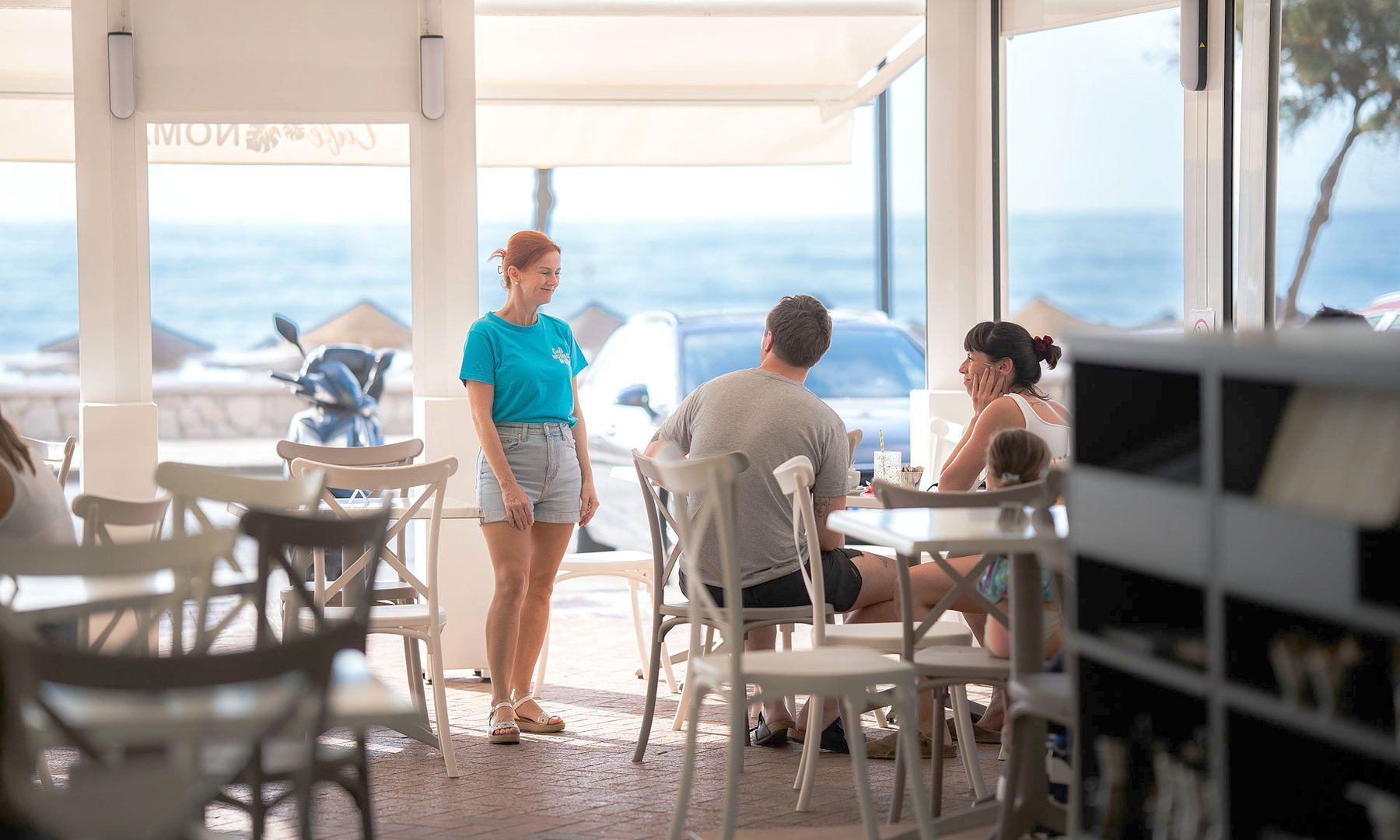 Café Nomad en servicio, las mesas frente a la playa y una camarera cruzando la sala.